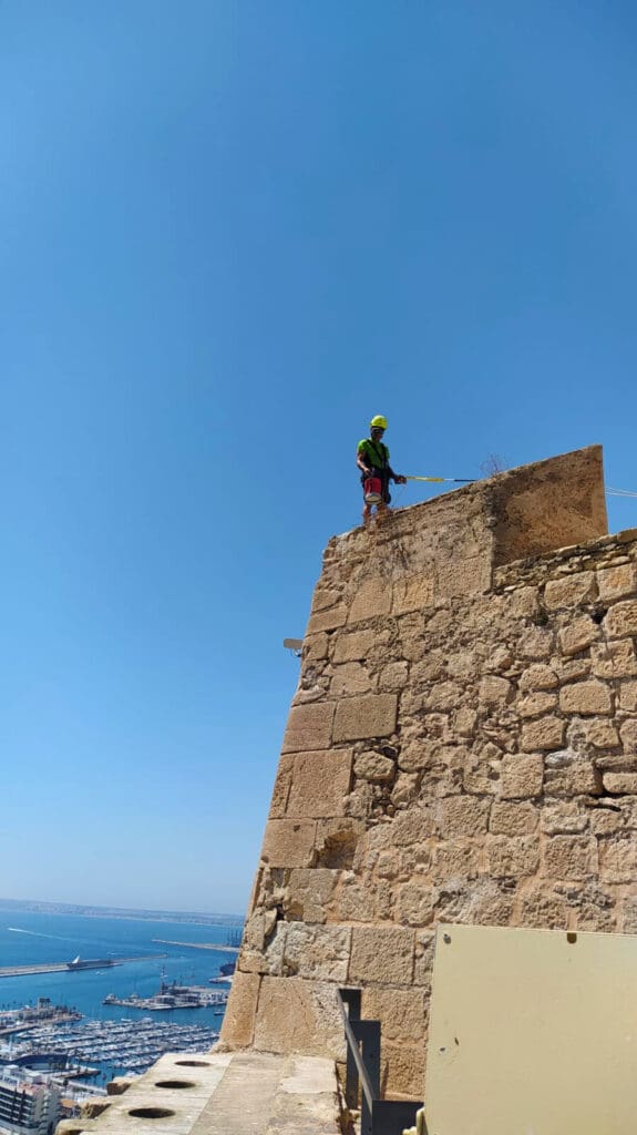 Operario de Verticales y Servicios realizando trabajos en la parte superior de la muralla del Castillo de Santa Bárbara, con vistas al puerto de Alicante.