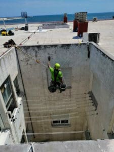 Operario de 'Verticales y Servicios', trabajando en la rehabilitación de un patio de luces. Está asegurado con arneses y cuerdas en la parte superior del edificio, pintando la pared exterior con vistas al mar de fondo.