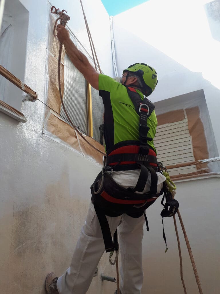 operario de 'Verticales y Servicios', trabajando en la pintura de un patio interior. El trabajador está asegurado con un arnés de seguridad y cuerdas mientras aplica pintura blanca en la pared. Se observa que las ventanas y otras áreas han sido cuidadosamente cubiertas con papel y cinta adhesiva para protegerlas de la pintura.