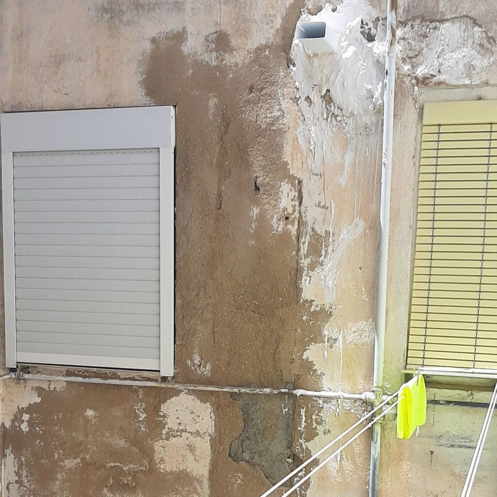 Pared de un patio de luces con evidentes signos de deterioro. Hay manchas de humedad y desprendimientos en el revestimiento. Dos ventanas con persianas están presentes, una blanca y otra amarilla. En la pared se observan grietas y parches de reparación. Esta imagen resalta la necesidad de mantenimiento y rehabilitación para mejorar la apariencia y la estructura del edificio.