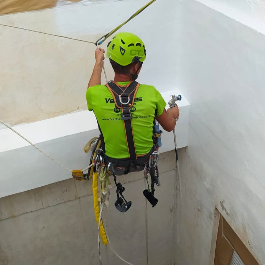operario de 'Verticales y Servicios', realizando trabajos de pintura en un patio de luces. Está asegurado con un arnés de seguridad y cuerdas mientras aplica pintura blanca en la pared. Se observa que las ventanas y áreas circundantes están protegidas con papel para evitar manchas.