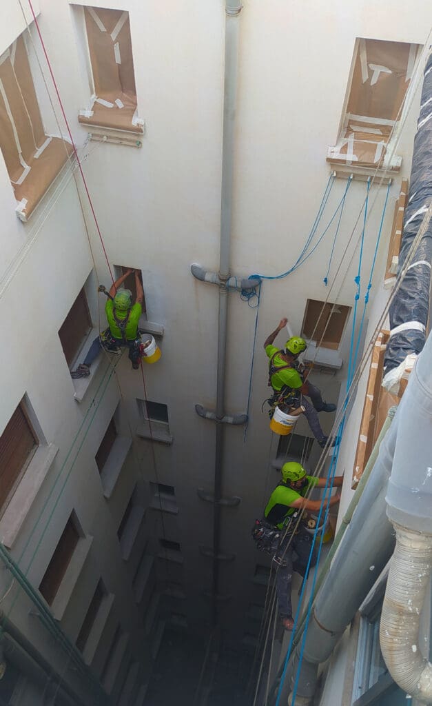 tres operarios de 'Verticales y Servicios' con cascos verdes y camisetas verdes, trabajando en la pintura de un patio de luces. Están asegurados con cuerdas y arneses de seguridad, mientras se elevan a distintas alturas. Las ventanas están cubiertas con papel y cinta adhesiva para protegerlas.