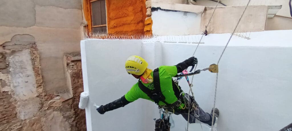 Operario de 'Verticales y Servicios' con un casco amarillo y una camiseta verde, trabajando en la pintura de un patio interior. El trabajador está asegurado con cuerdas y un arnés de seguridad mientras aplica pintura blanca en las paredes.
