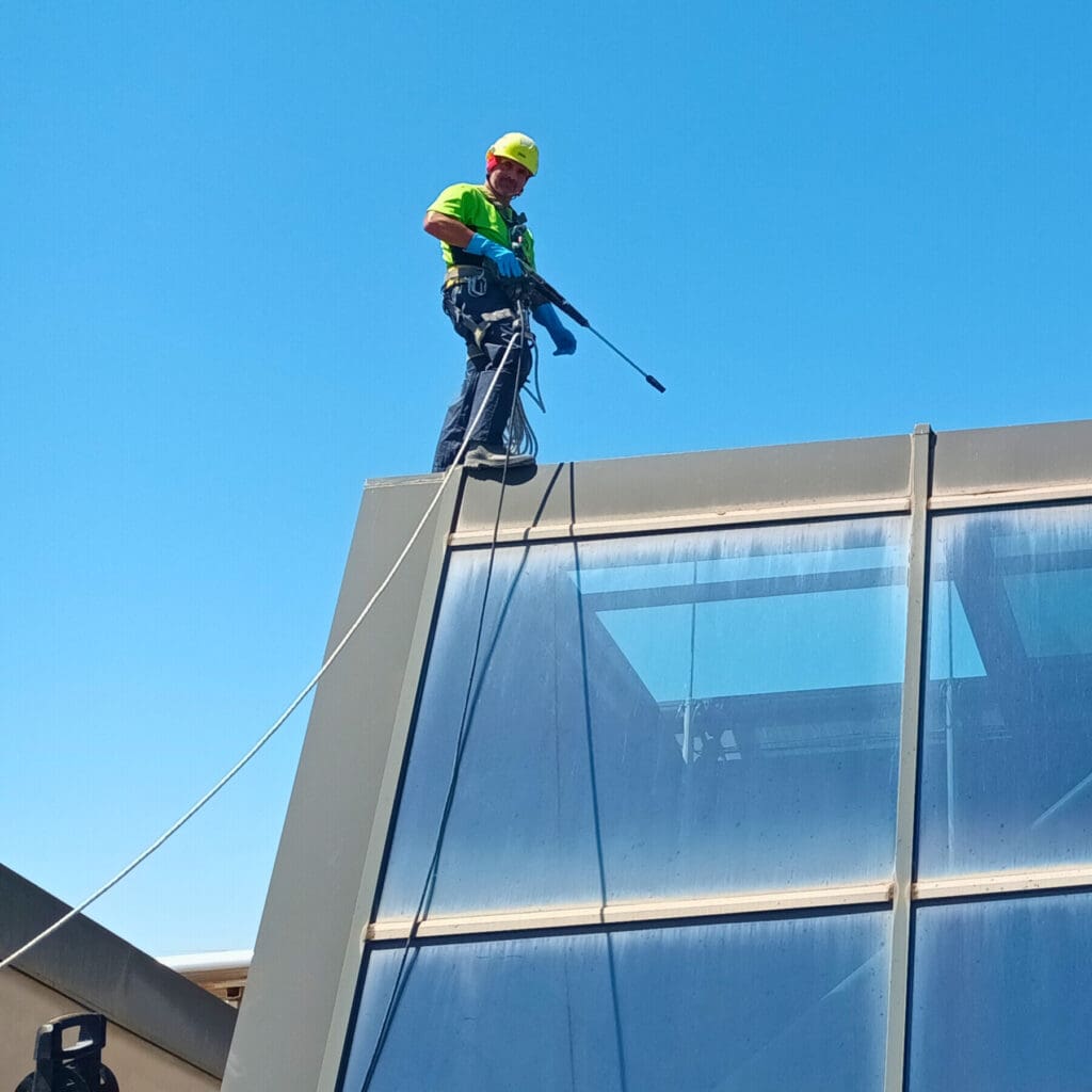 Empleado de 'Verticales y Servicios' trabajando en altura para limpiar los cristales de un centro comercial en Murcia, usando equipo de seguridad y una hidrolimpiadora.