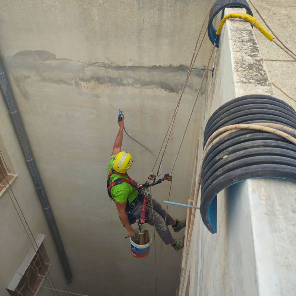 Trabajador vertical de 'Verticales y Servicios' con un casco amarillo y una camiseta verde, realizando trabajos de pintura en altura en un patio de luces. El trabajador está asegurado con un arnés de seguridad y cuerdas mientras aplica pintura con una pistola pulverizadora. Lleva un cubo de pintura colgado de su arnés. La pared en la que trabaja muestra signos de humedad y desgaste, lo que resalta la necesidad de rehabilitación.