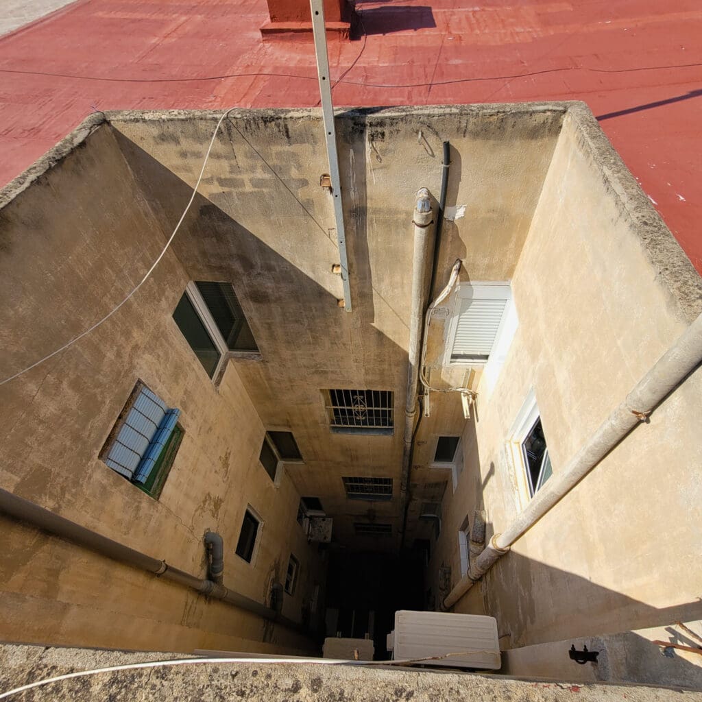 La foto muestra un patio de luces de un edificio visto desde la parte superior. Las paredes del patio están desgastadas y necesitan mantenimiento, con manchas de humedad y grietas visibles. Hay varias ventanas que dan al patio, algunas con rejas y otras con persianas. También se observan tuberías verticales que recorren las paredes y equipos de aire acondicionado instalados. El techo del edificio es de color rojo y tiene algunas estructuras adicionales visibles en la parte superior. La imagen ilustra la necesidad de una rehabilitación para mejorar la estética y funcionalidad del espacio.
